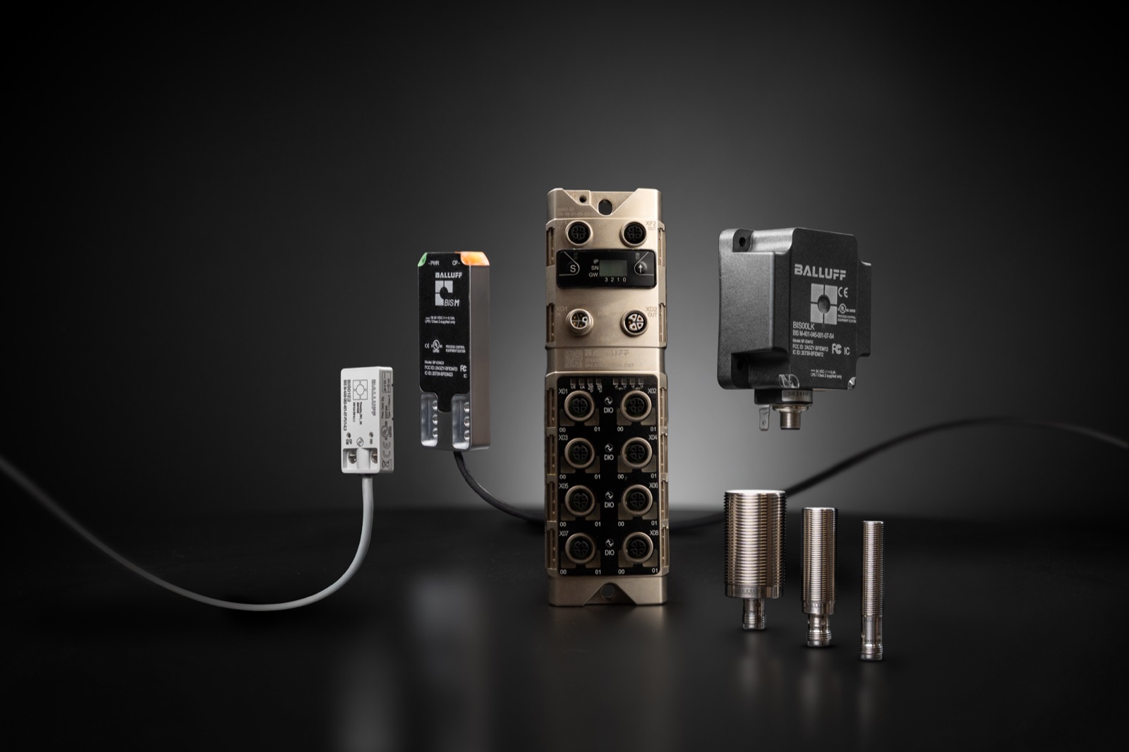Balluff IO-Link family on a dark studio background: an IO-Link master block with eight M12 ports, a BIS M unit, an RFID read head, and sensors.